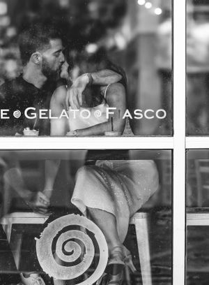Black and white photo through window showing someone sitting at a cafe with 'Gelato' text and spiral design visible.  Brunswick, Maine.