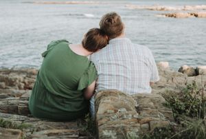 Two people in casual clothing sitting close together on coastal rocks while looking out at the ocean view.