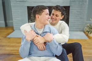 Two people in cozy white sweaters share a tender embrace while sitting on wooden floor in front of a fireplace.  Taken in Bath, Maine.