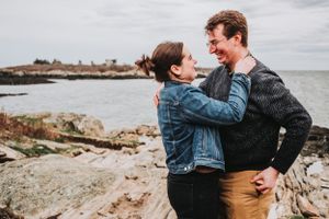A couple in denim jacket and sweater embrace lovingly on rocky coastal shore during overcast day.