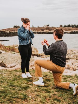 A romantic proposal moment unfolds on rocky coastal shores with water and autumn foliage in the background.  Land's End on Bailey Island, ME