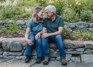 A couple sits on a rustic stone wall in a garden, dressed casually in denim and shown from the legs down.
