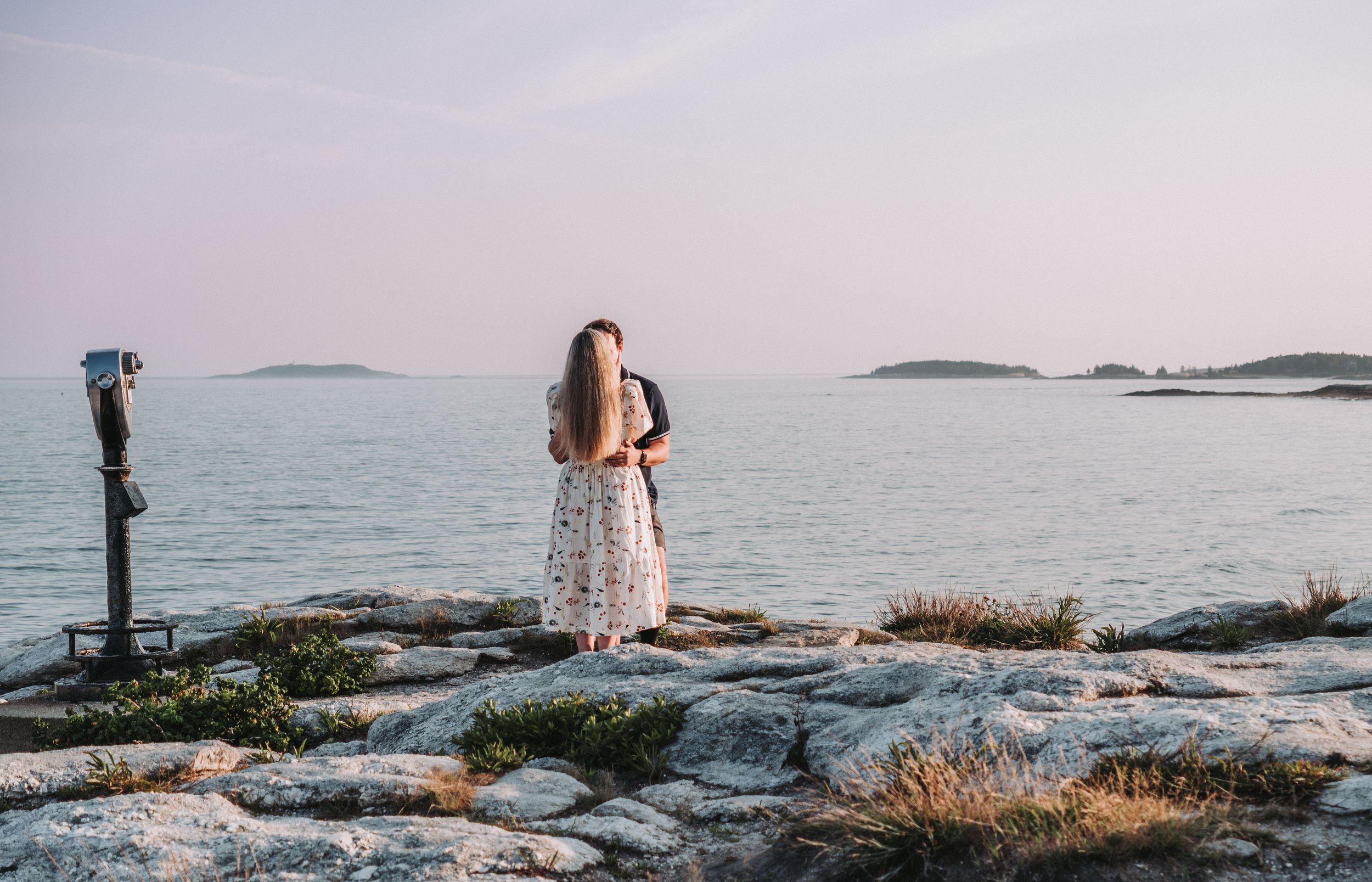 A coastal landscape photos shows a neon pink sunset skies over rocky shores and calm ocean waters.  A newly engaged couple embraces.