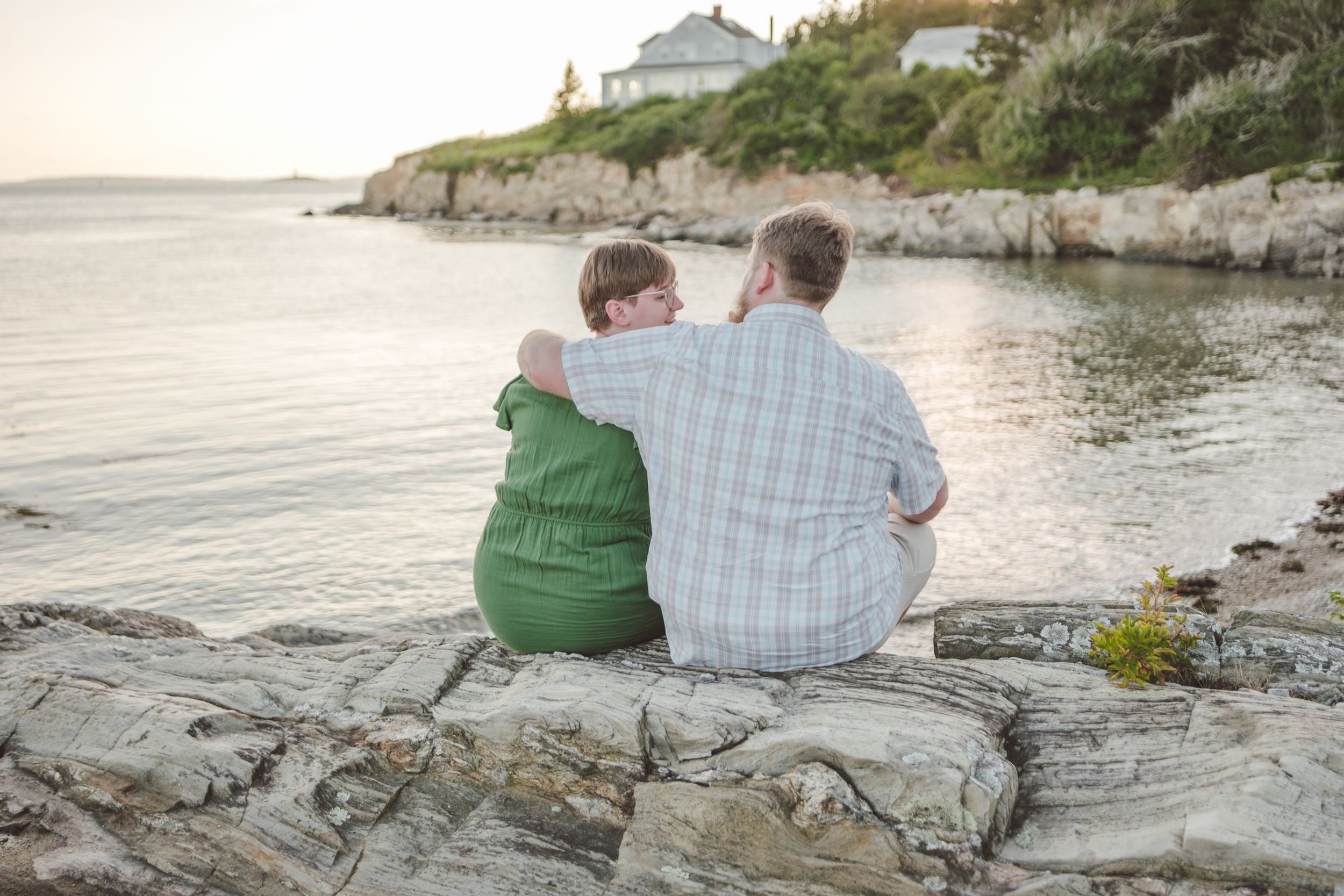 Two people sit on coastal rocks at sunset with ocean views and beach houses in the background.  Shot at Land's End in Bailey Island, ME.
