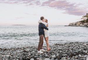 Two people embrace on the rocky beach at Fort Williams Park in Cape Elizabeth, Maine at sunset with the ocean and cliffs in the backdrop.