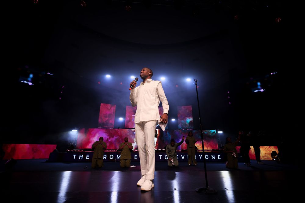 Artist in white suit performing on dimly lit stage with red accents and venue signage.