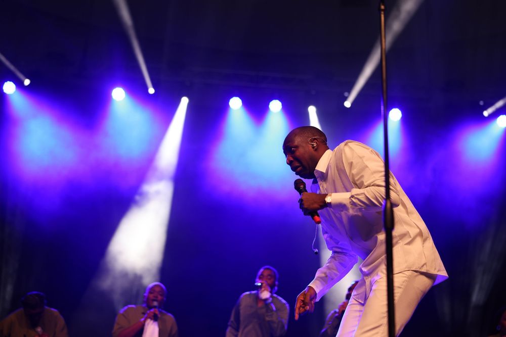 Performer in white attire energetically singing on stage with dramatic blue lighting and backup performers.