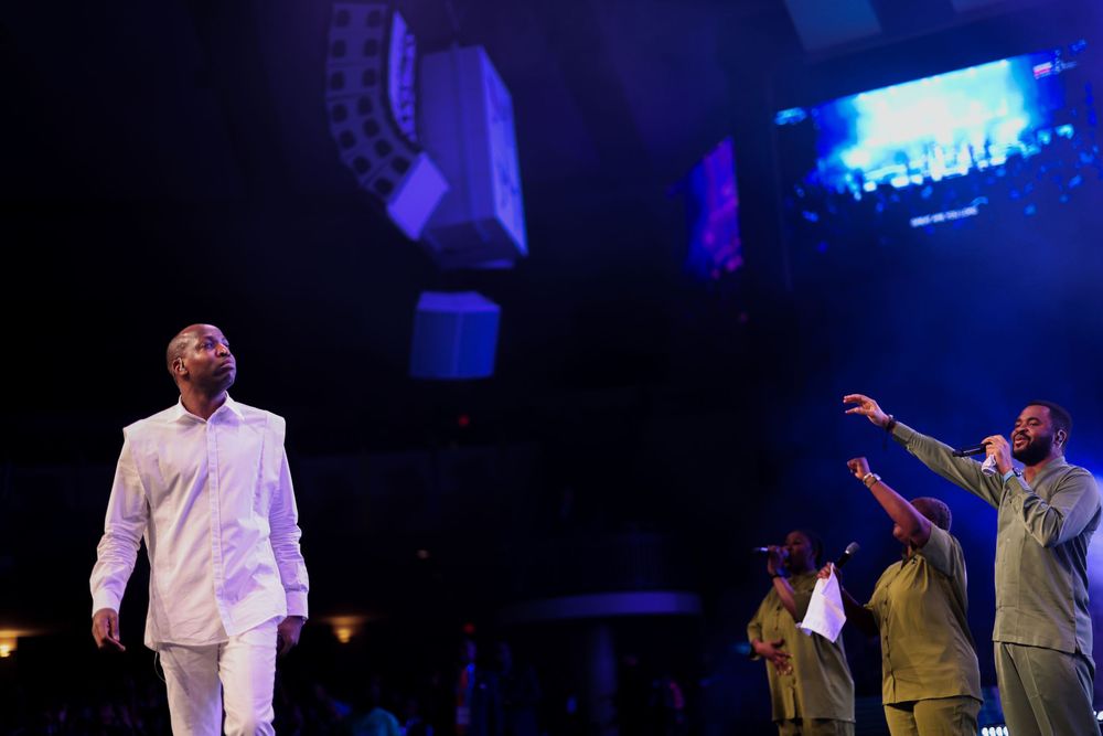 Performers in white outfits on stage with dramatic lighting and audience silhouettes.