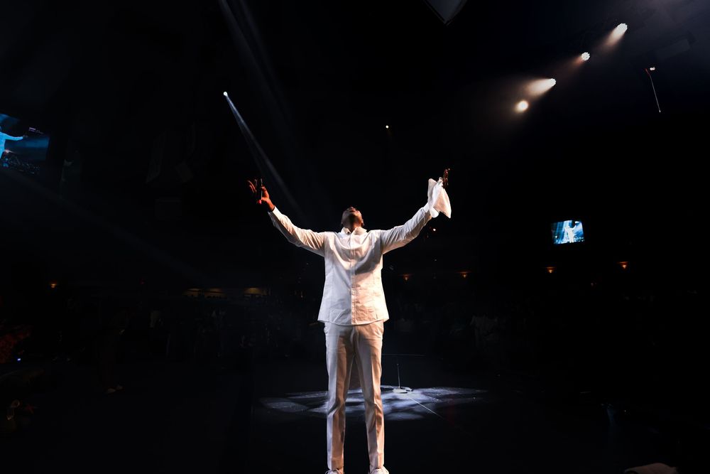 Performer in white suit strikes triumphant pose under spotlight on dark stage.