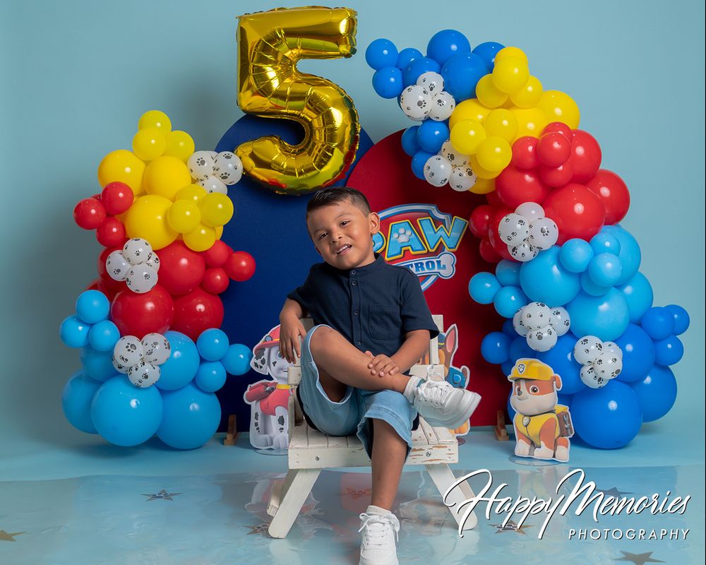 A young boy celebrates his 5th birthday with a colorful Paw Patrol balloon arch backdrop in red, blue, and yellow.