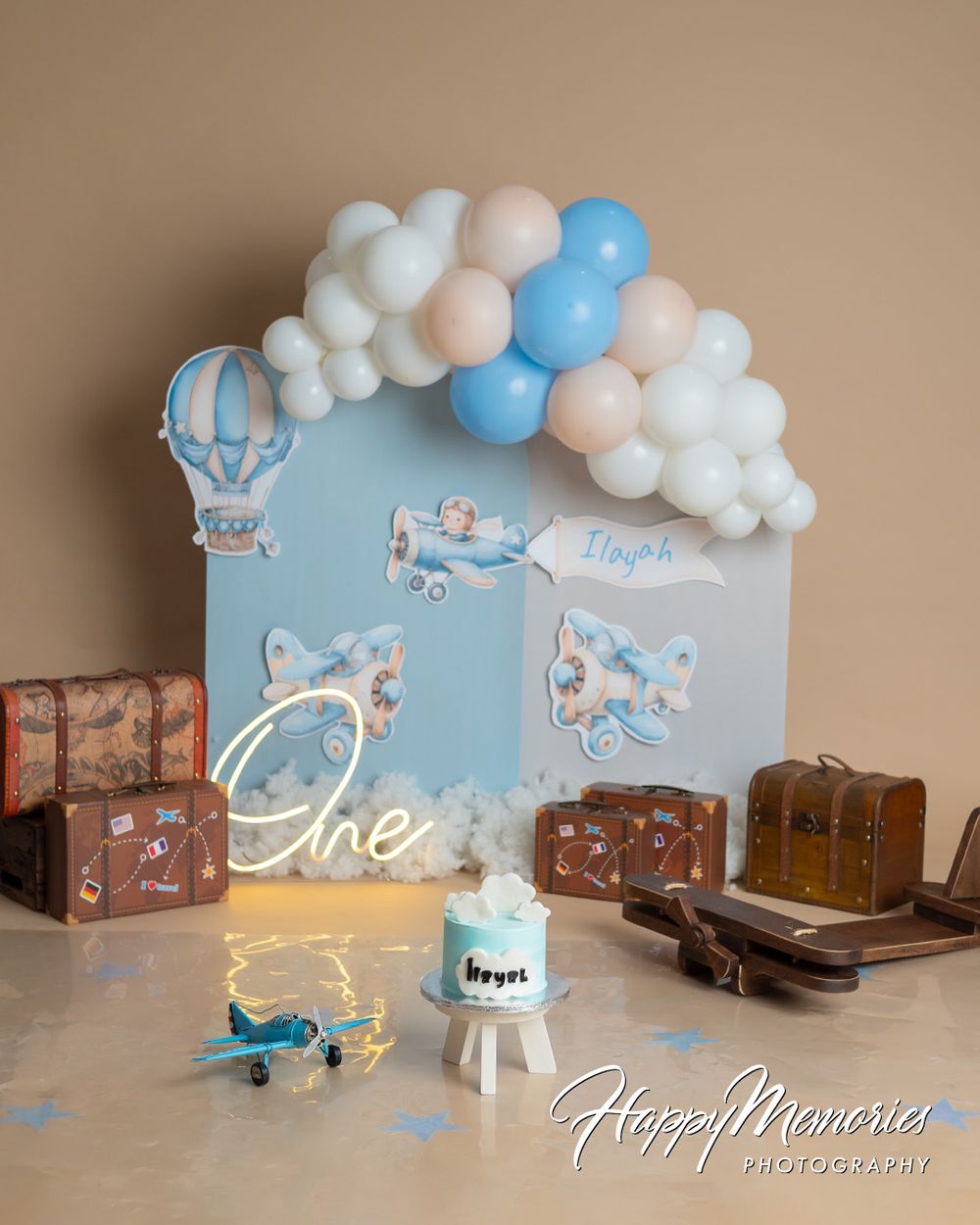 First birthday cake smash setup with airplane theme, balloon arch, vintage suitcases, and personalized blue backdrop.
