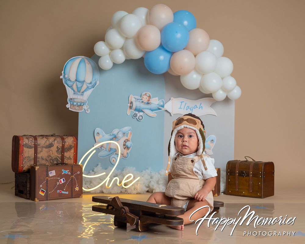 Baby's first birthday cake smash photo with balloon arch, vintage suitcases, and aviator hat in blue and white studio.