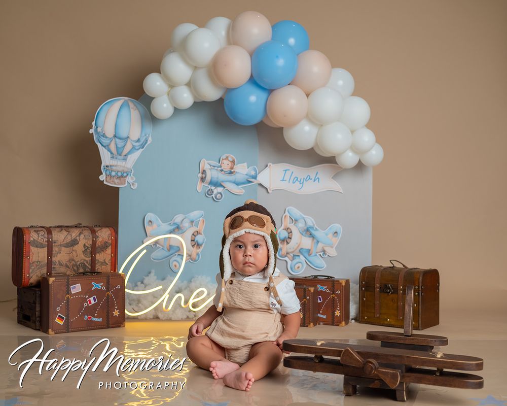 Baby's first birthday cake smash photo with aviator hat, balloon arch, vintage suitcases, and travel theme backdrop.