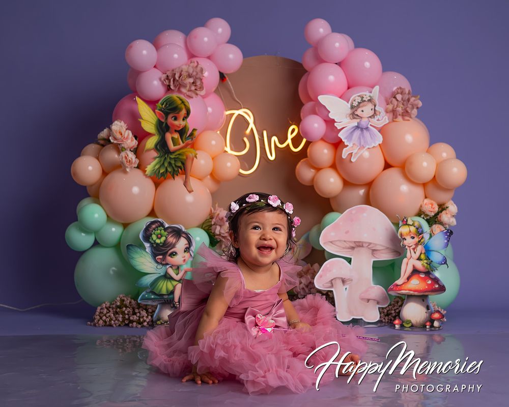 Baby girl in pink tutu celebrates first birthday with fairy-themed balloon arch backdrop reading One.