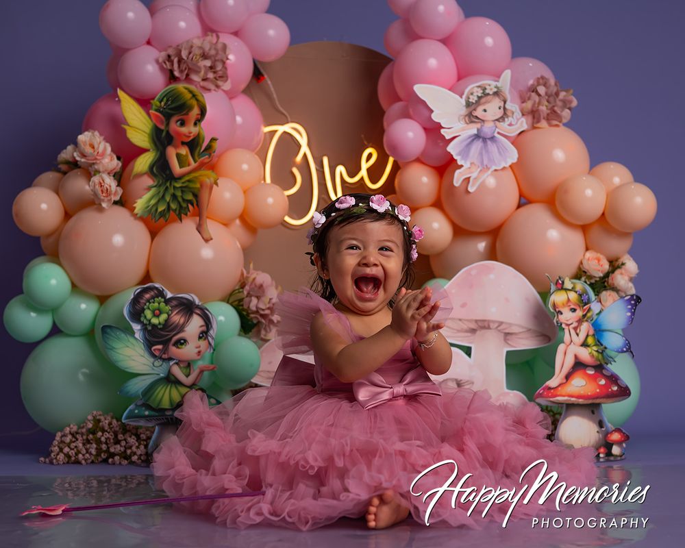 Joyful baby girl in pink tutu celebrates first birthday with fairy-themed balloon backdrop and neon “One” sign.