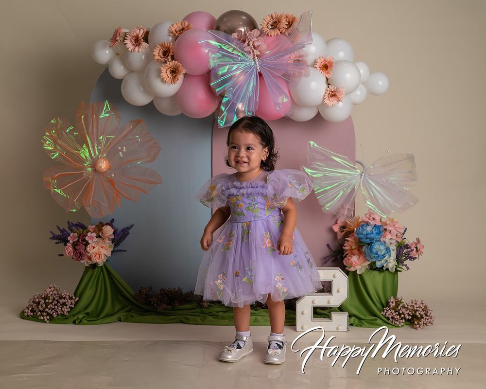 Toddler girl in purple tutu dress poses at fairy-themed 2nd birthday cake smash setup with balloons and flowers.