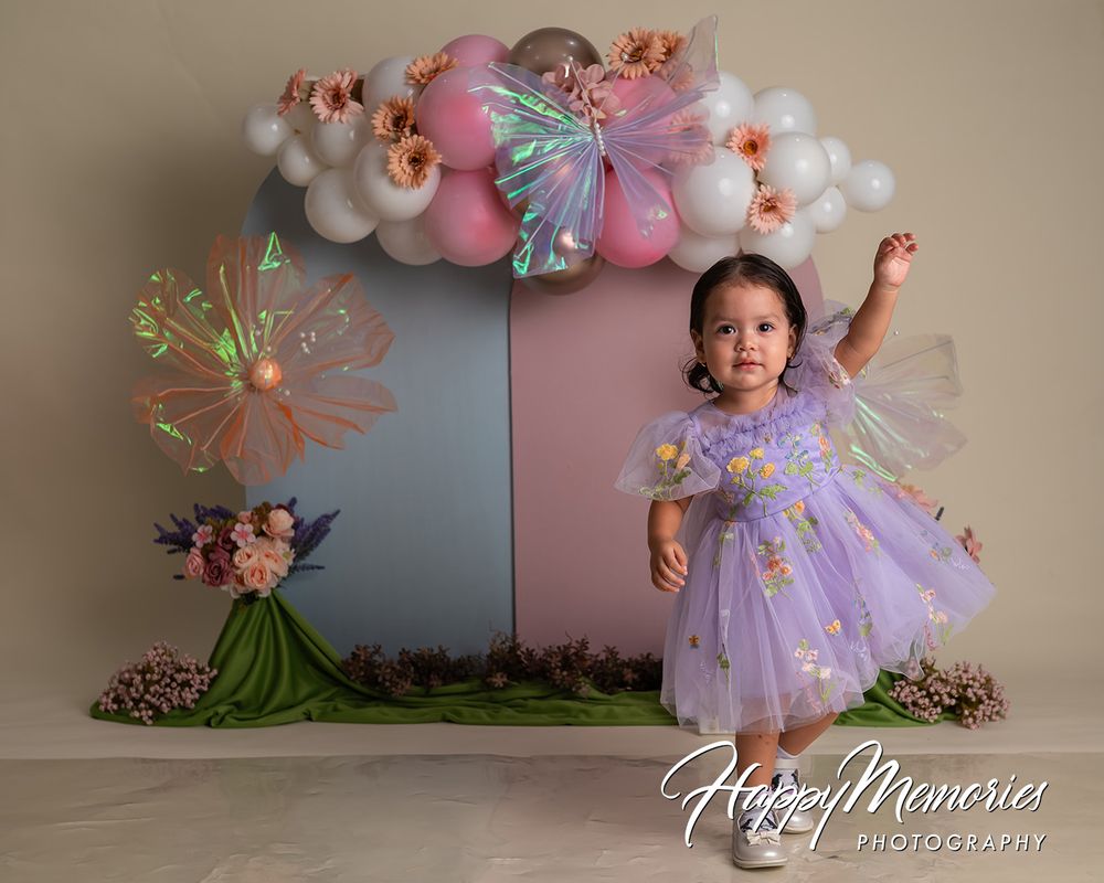 Toddler girl in purple tutu dress poses before balloon arch backdrop at first birthday cake smash photo session.