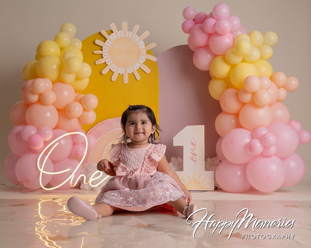 A baby girl in a pink dress sits before a colorful balloon arch and sun backdrop at a first birthday photoshoot.