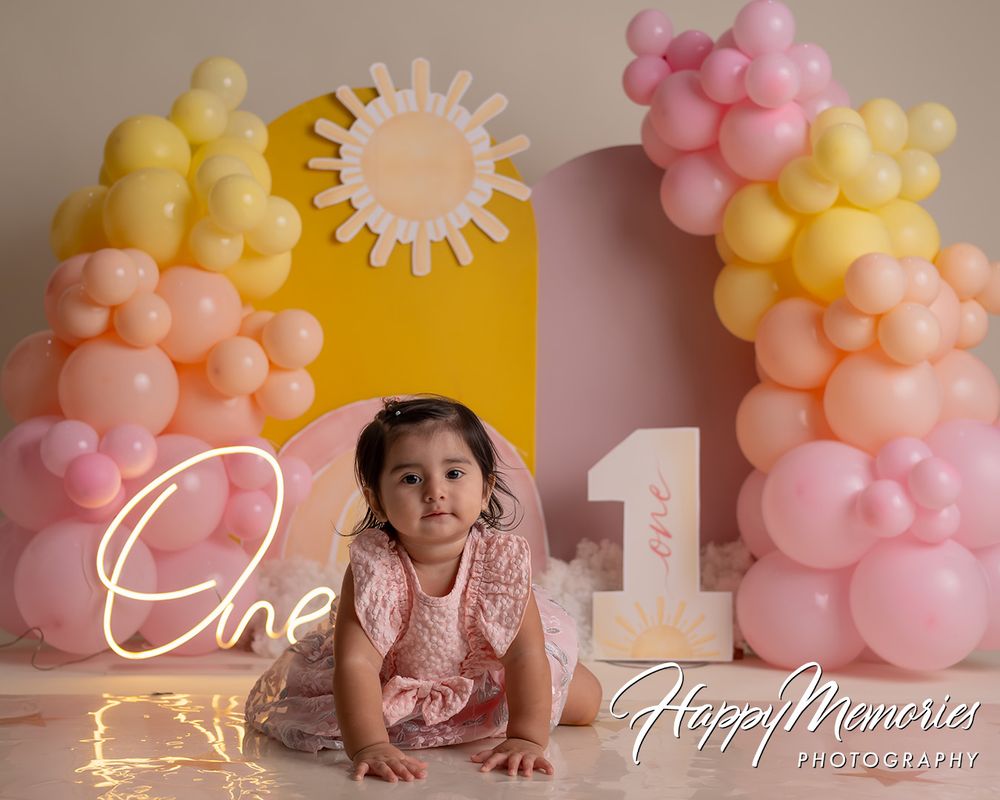 Baby girl in pink dress poses before colorful balloon arch and sun backdrop for first birthday photo.