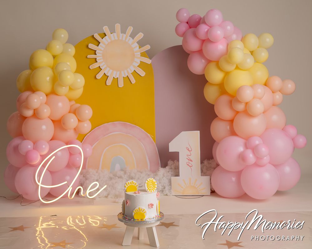 First birthday cake smash setup with pink, yellow, and peach balloon arch, rainbow decor, and sun backdrop.