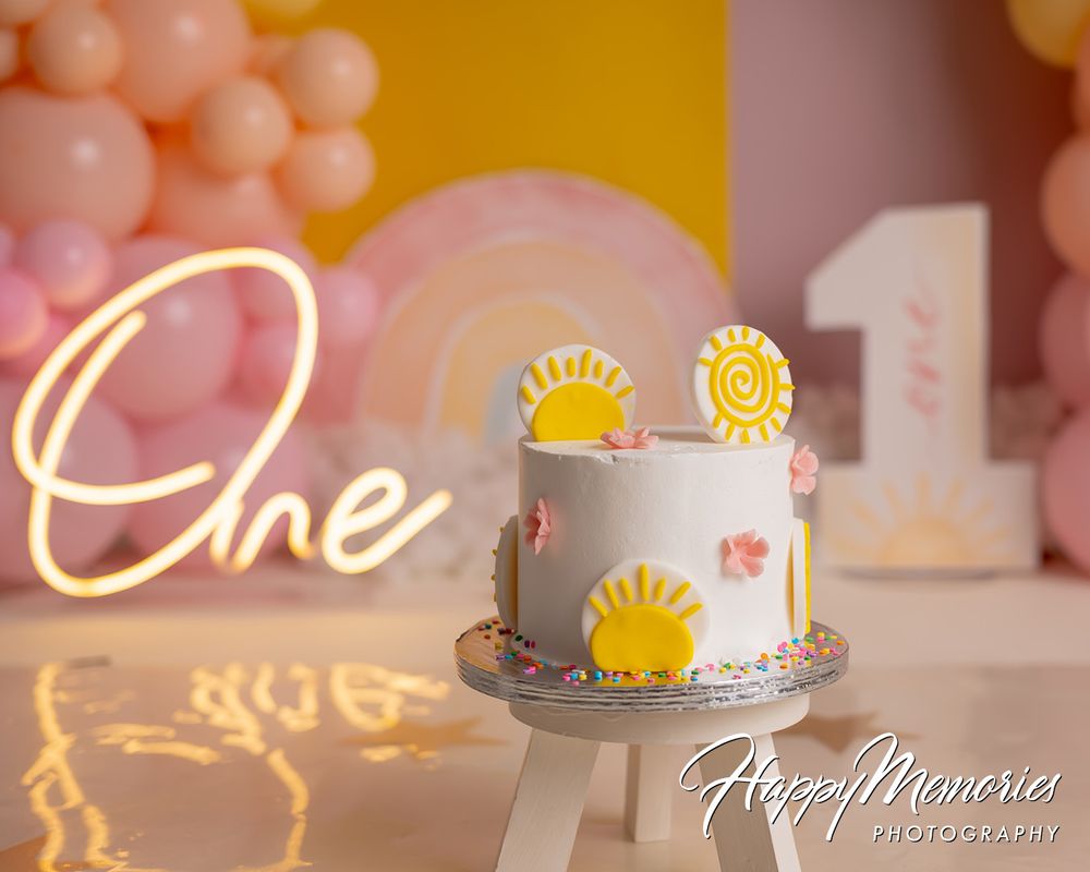 White first birthday cake with yellow sun decorations, pink balloons, and glowing One sign for a sunshine-themed party.