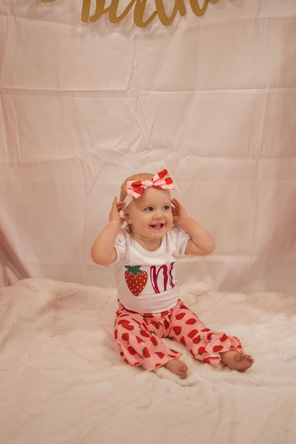 Baby wearing strawberry-themed outfit sits on white backdrop during photo session.