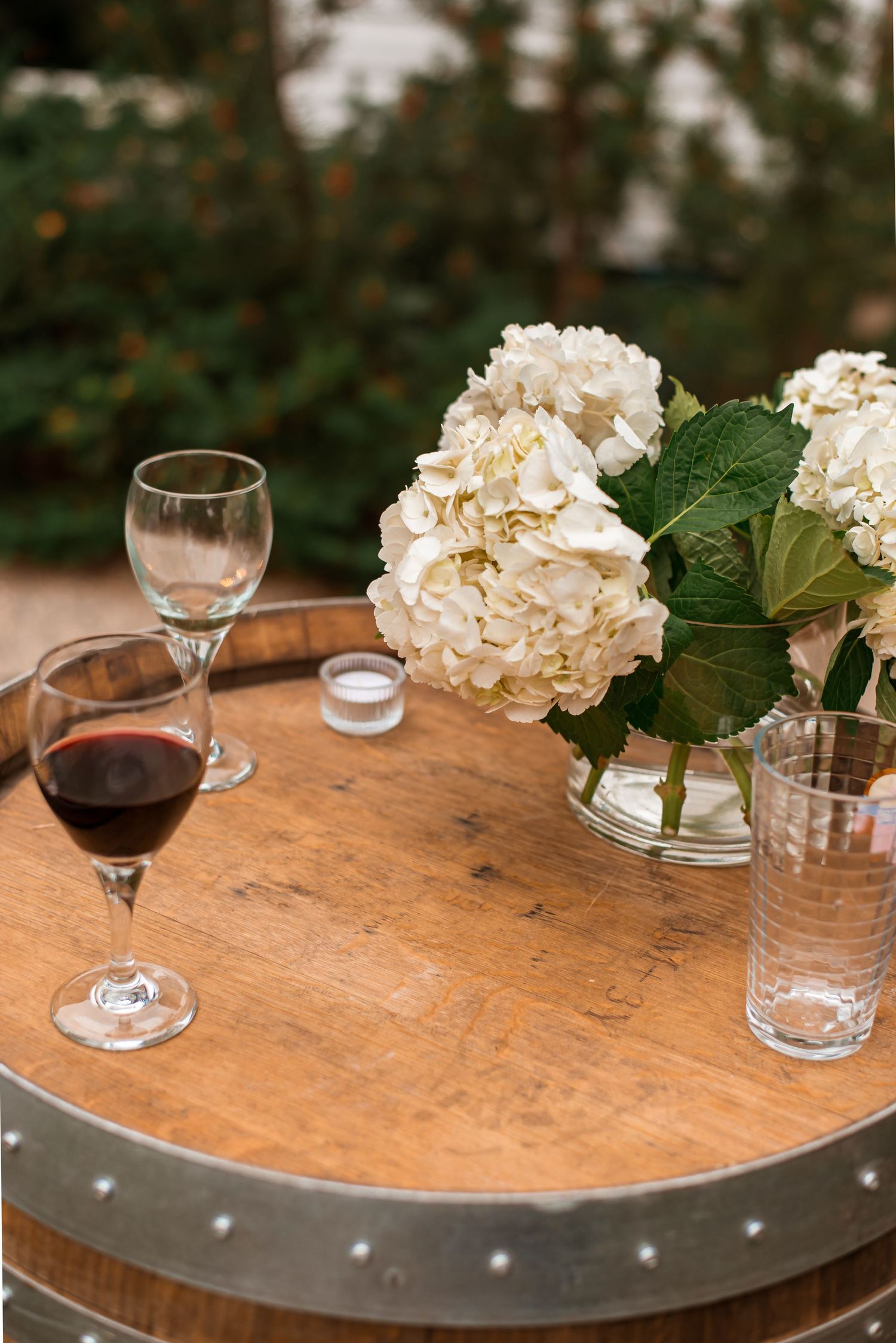 White hydrangeas in a glass vase sit beside wine glasses on a rustic wooden barrel table.