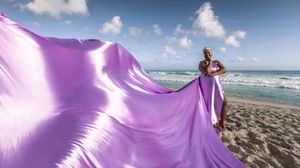 Purple fabric flows gracefully in the wind against a scenic beach backdrop with blue ocean waves and cloudy sky.