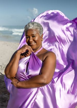 A person in a flowing purple dress poses expressively against a beach backdrop at sunset.