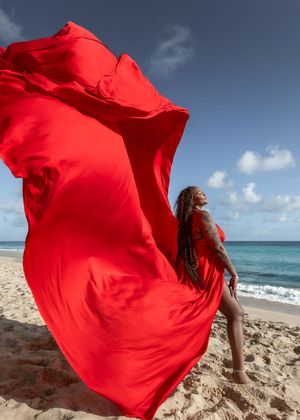 Red fabric billows dramatically against a beach backdrop with turquoise ocean waters and cloudy sky.