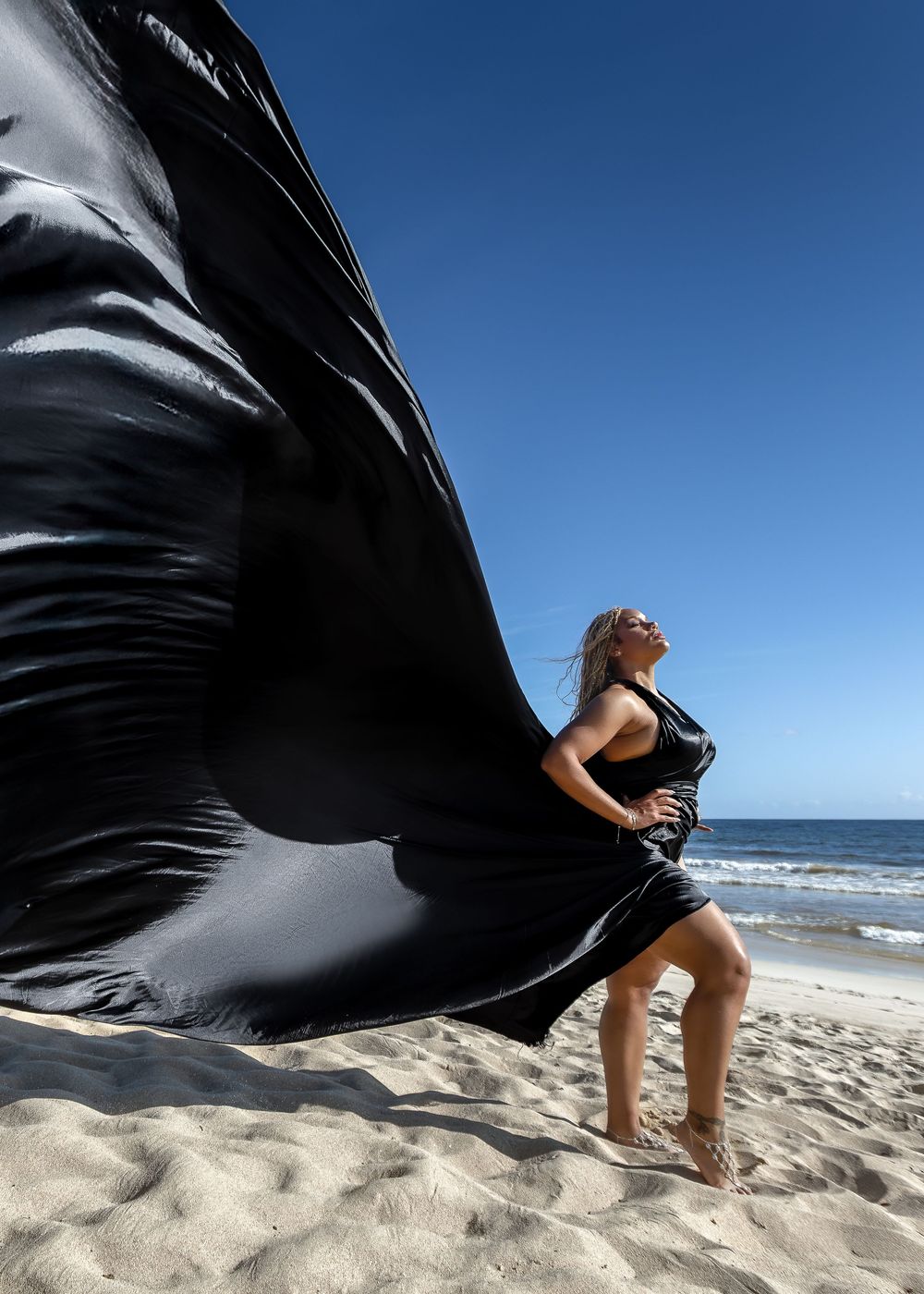 A flowing black dress billows dramatically in the wind against a beach backdrop with blue sky and ocean.