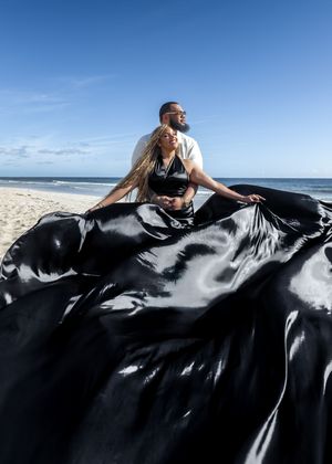 A person in black clothing lying on a sandy beach with flowing dark fabric billowing in the ocean breeze.