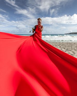 A flowing red dress billows dramatically against a beach landscape with blue sky and ocean waves in the background.
