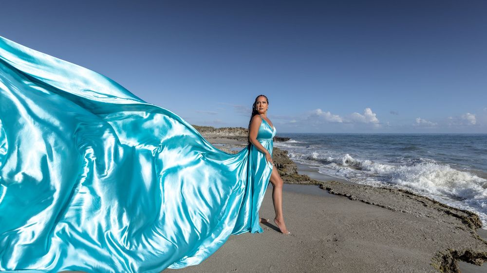 A flowing turquoise fabric billows gracefully against a beach landscape with crashing waves and blue sky.