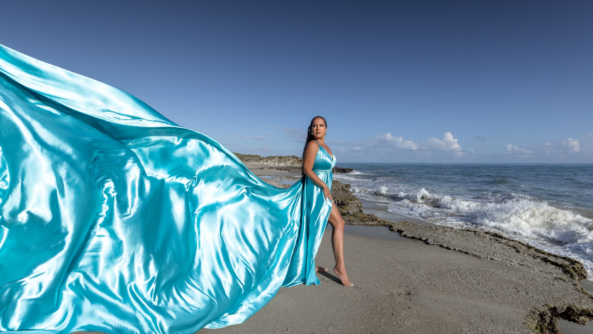 A flowing turquoise fabric billows gracefully against a beach landscape with crashing waves and blue sky.