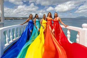 People wearing colorful rainbow dresses stand together on a boat deck against a bright blue sky and ocean view.