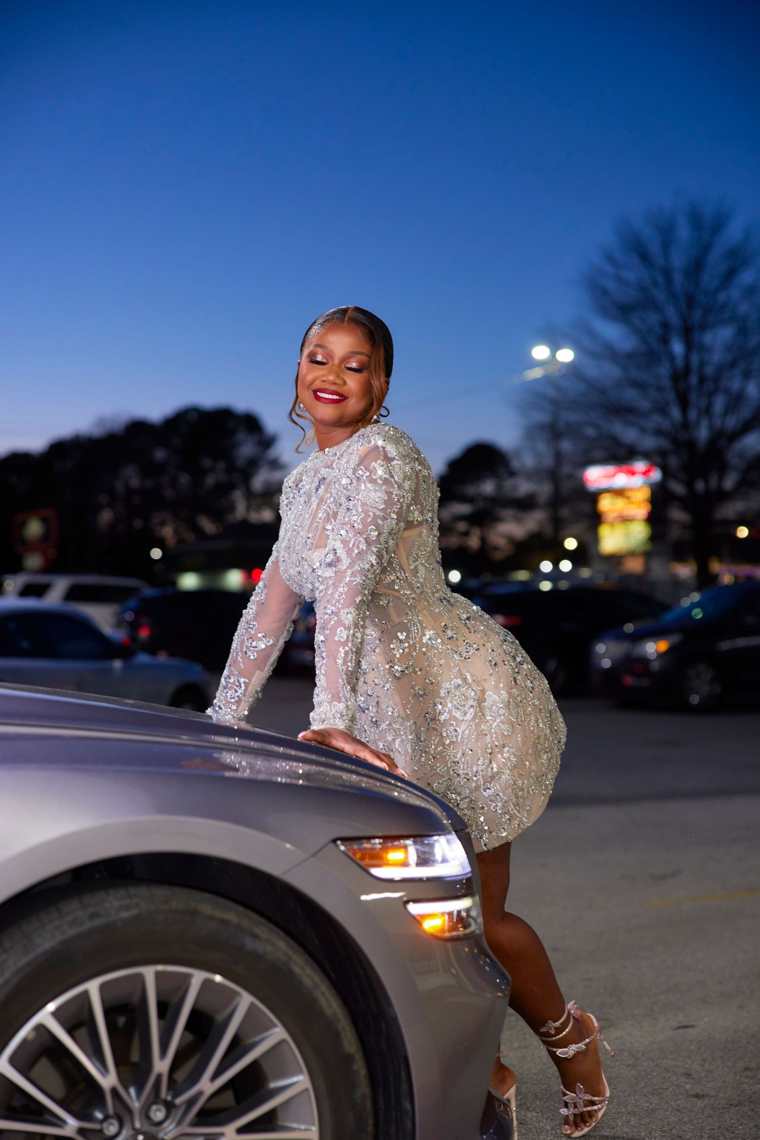 A sparkly sequined dress shimmers in an evening photoshoot against a car in a parking lot at dusk.