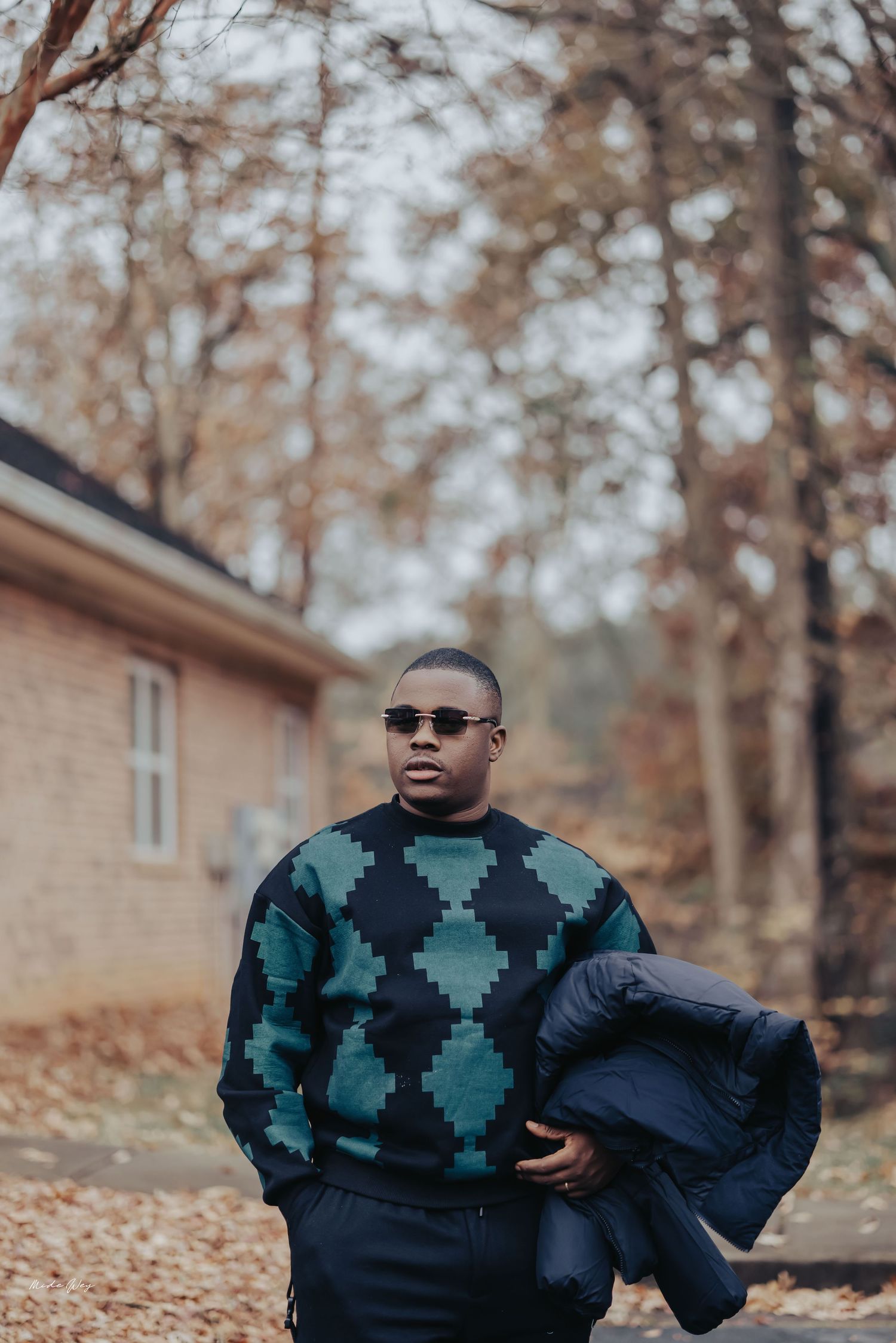 A person in sunglasses and a patterned sweater poses outdoors on fallen autumn leaves near a building.