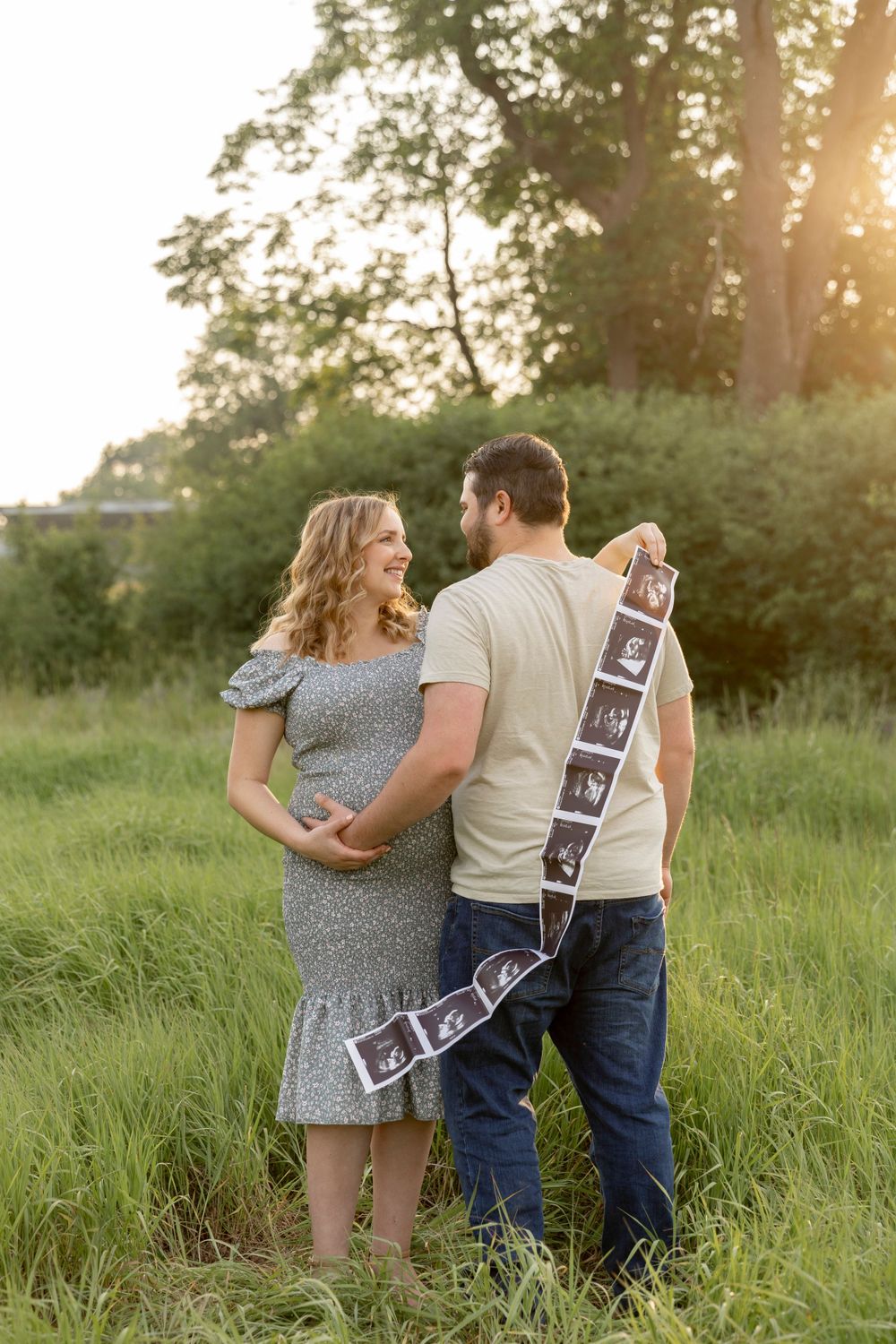 pregnant couple smiling at each other holding their ultrasound strip in a gray field in troy Michigan at sunset