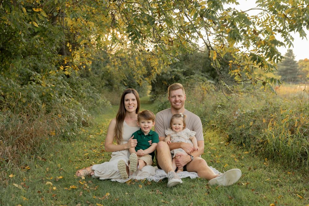 A family sit together on a blanket in a grassy field during golden hour while wearing coordinated neutral outfits at troy farm park michigan