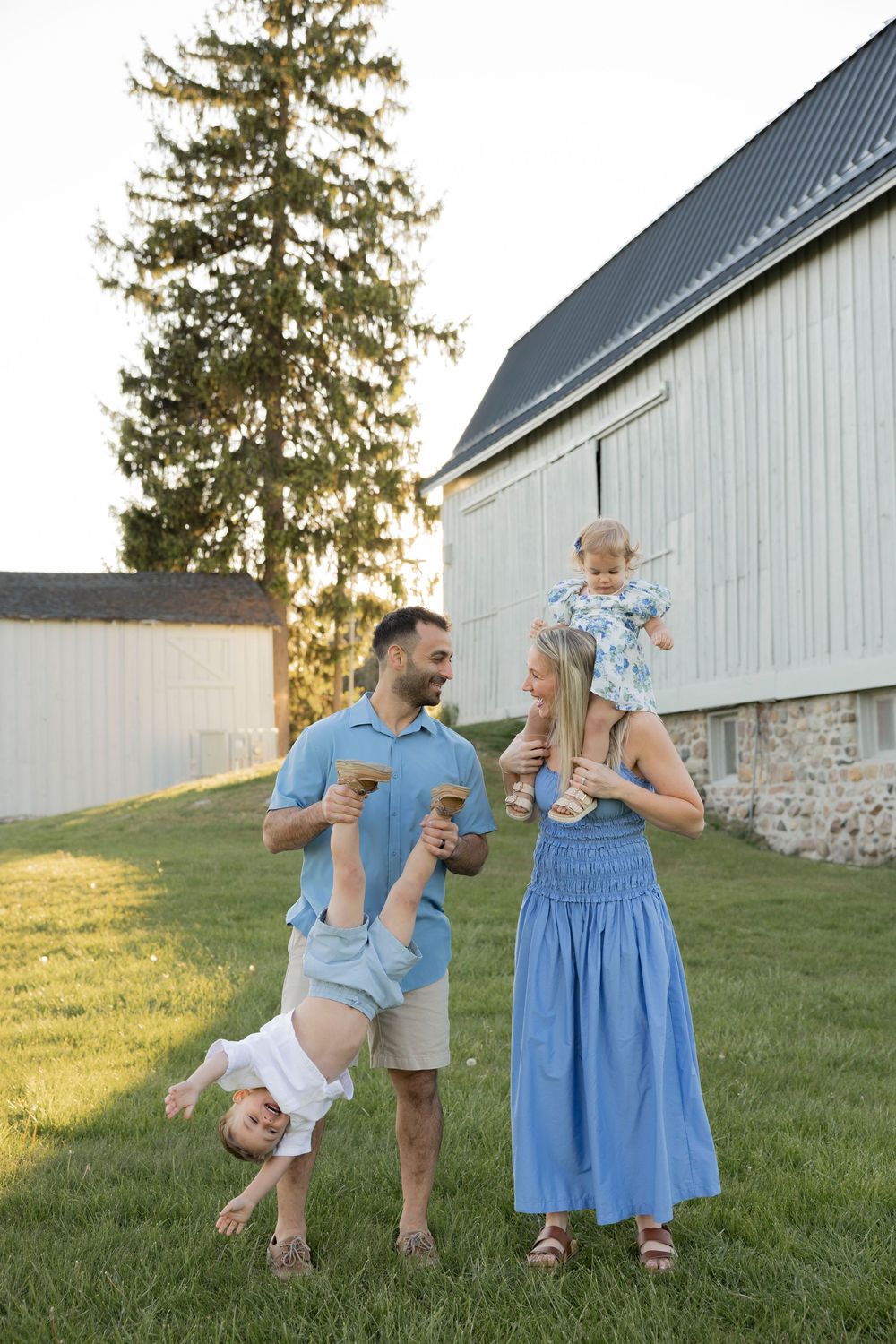 A family in blue dresses play near a white barn at troy farm park in michigan with dad playfully holding a toddler upside down.