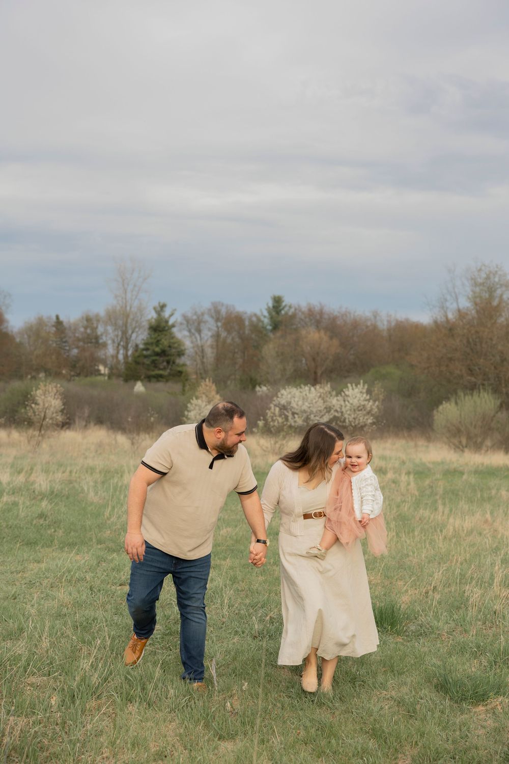 Family walking hand in hand through a grassy meadow at troy farm park in Michigan on a spring day with blooming trees in the background.