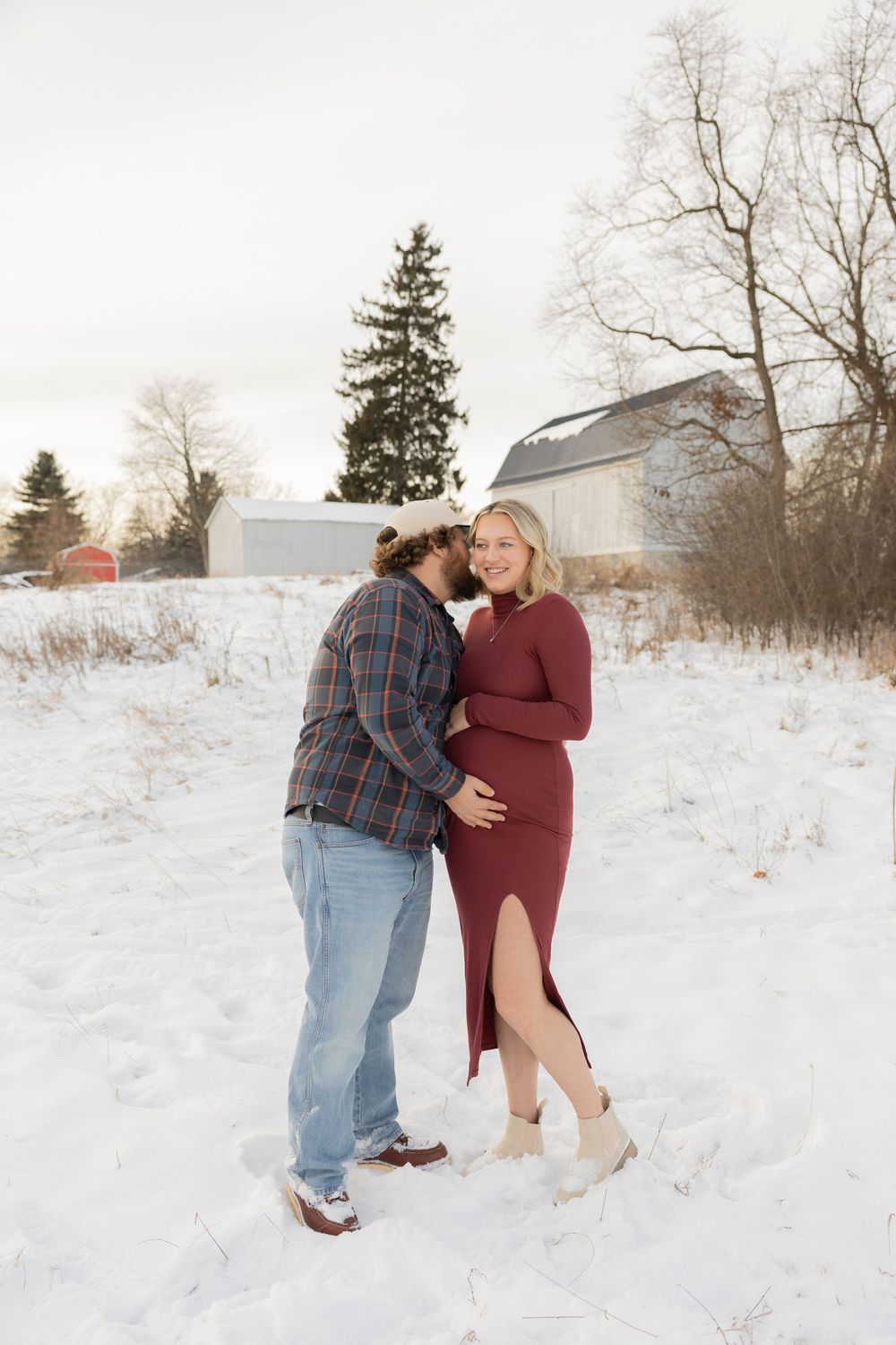A pregnant couple shares a romantic moment in a snowy winter landscape wearing burgundy dress and plaid shirt at troy farm park in michigan