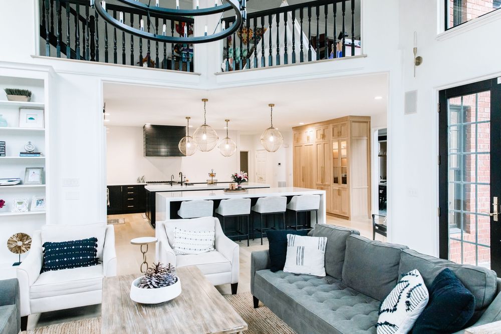 Open-concept living room and kitchen with gray sofa, white chairs, black cabinetry, and second-floor balcony.