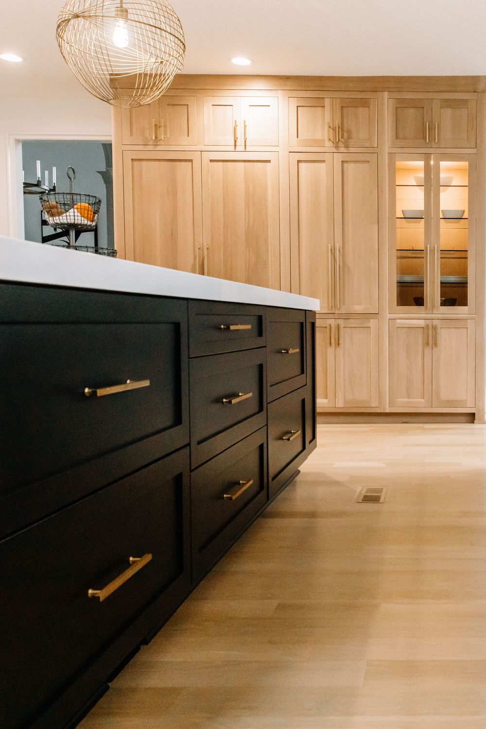 Black kitchen island with gold hardware contrasts light wood cabinetry and pale hardwood floors.