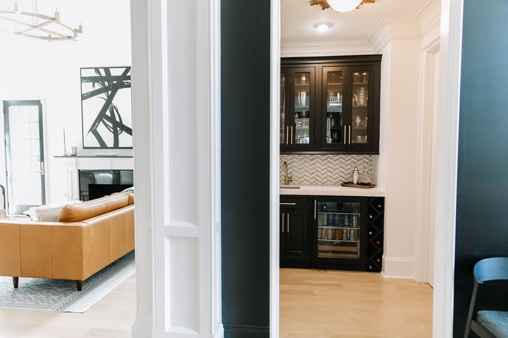 Dark butler pantry with glass cabinets, wine fridge, and herringbone tile backsplash viewed through doorway.