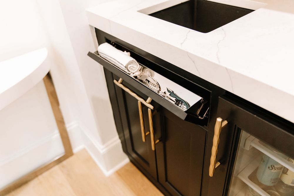 Black kitchen cabinet drawer with gold hardware pulls beneath white marble countertop near wine cooler.