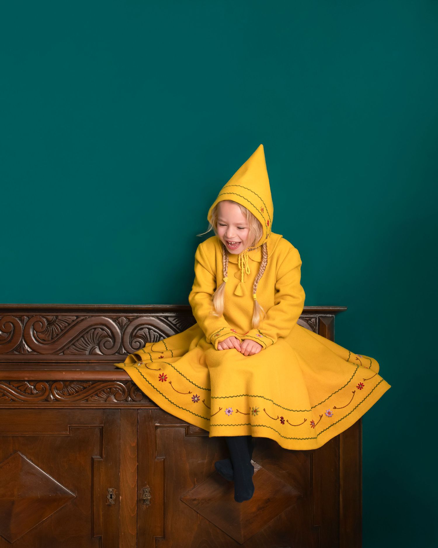 Wearing a yellow beautifully embroidered handmade dress and hat, child sits playfully on an antique wooden cabinet against a teal wall.