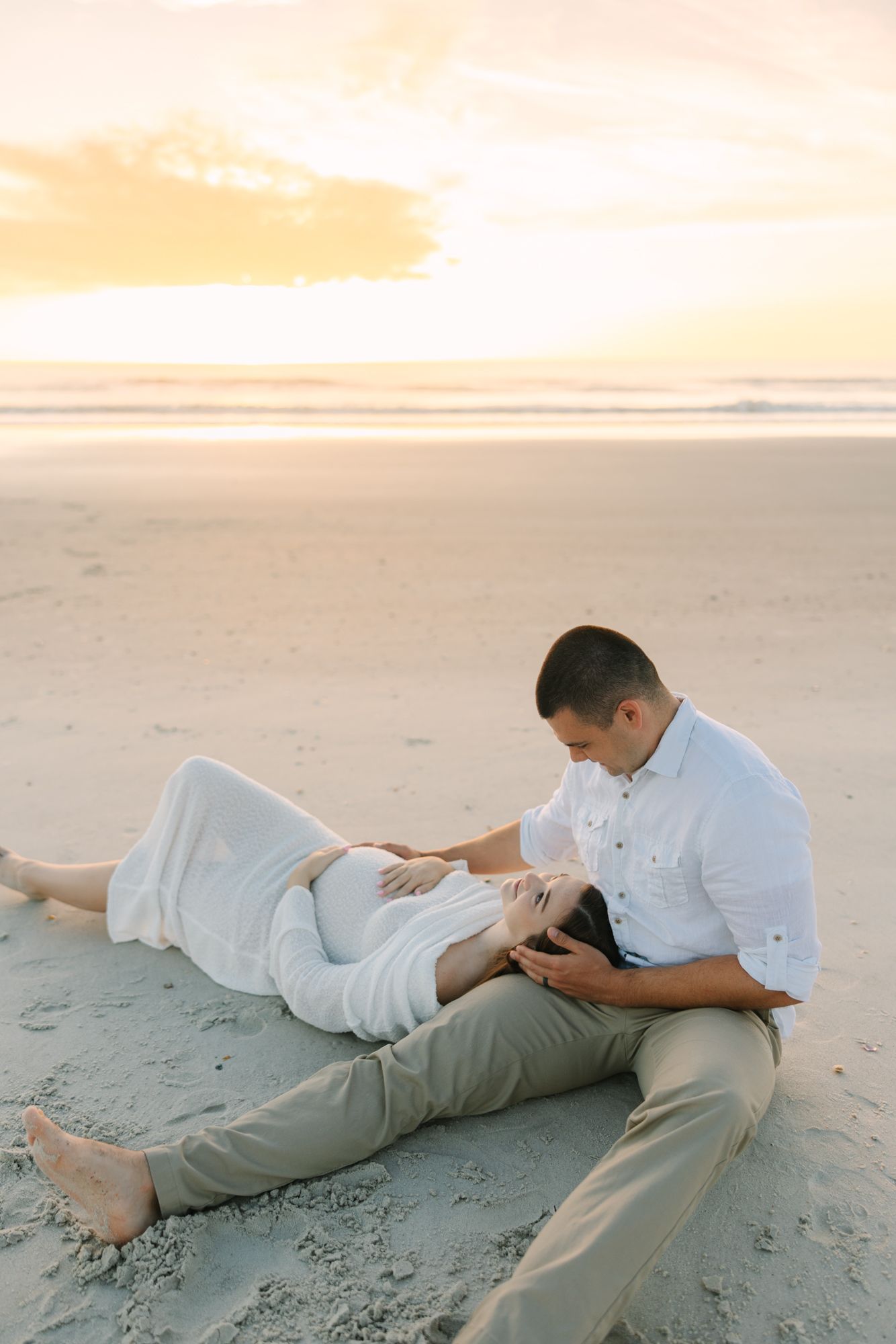 couple laying on the beach at sunrise in Jacksonville Beach, fl rubbing wife's pregnant belly