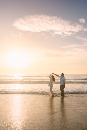 maternity couple at sunrise with sun coming over the ocean and man twirling woman